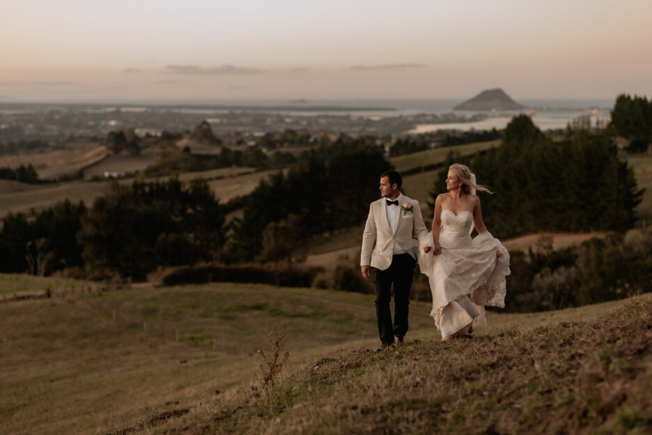 bride and groom walking in field with Mount Maunganui behind them