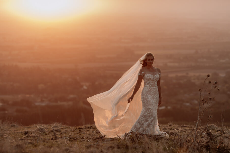 Bride standing on hill at sunset with her veil flying out
