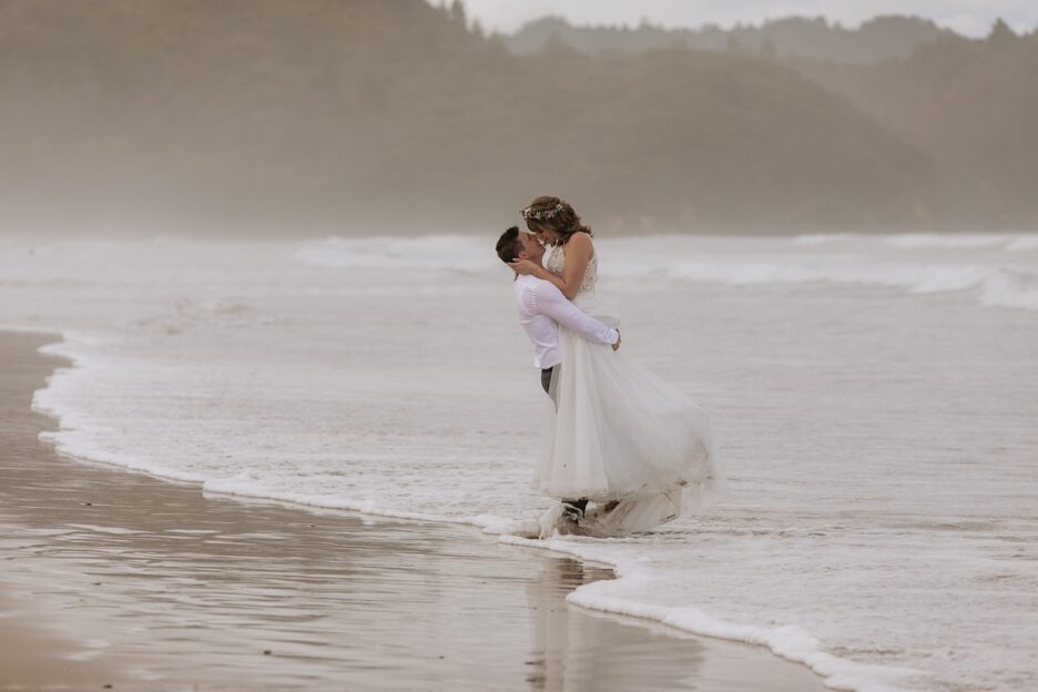 Waihi Beach Wedding groom lifting bride and kissing as waves crash around them