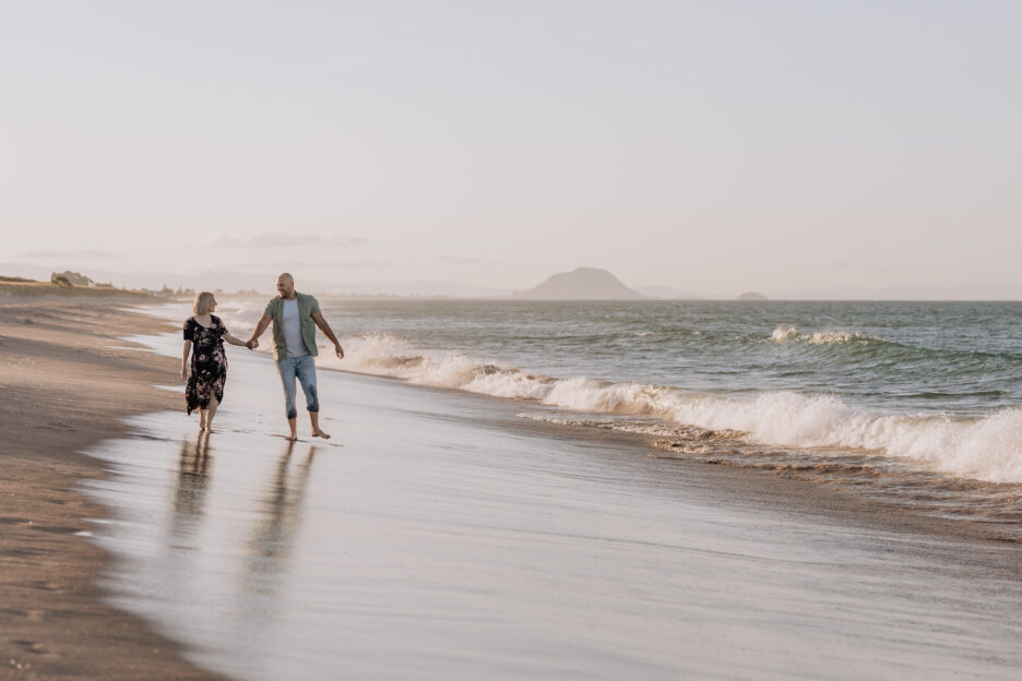 Lifestyle maternity photo of mum and dad walking on the beach waves crashing