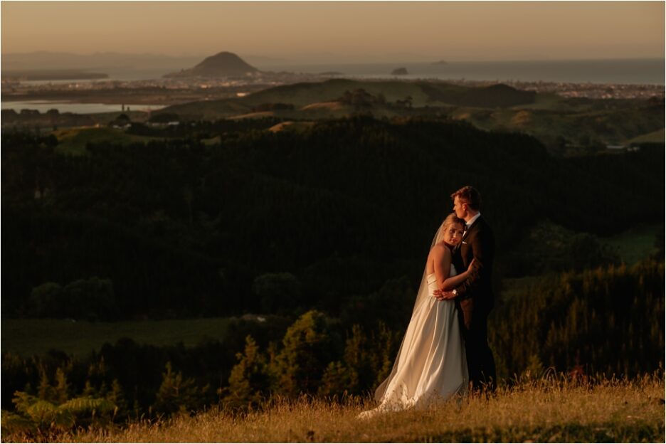 Beautiful sunset photo of bride and groom on hills in New Zealand with Mount Maunganui behind them