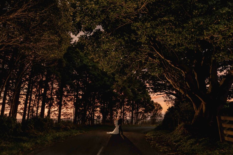 Photo of bride and groom on road with trees lit up at sunset