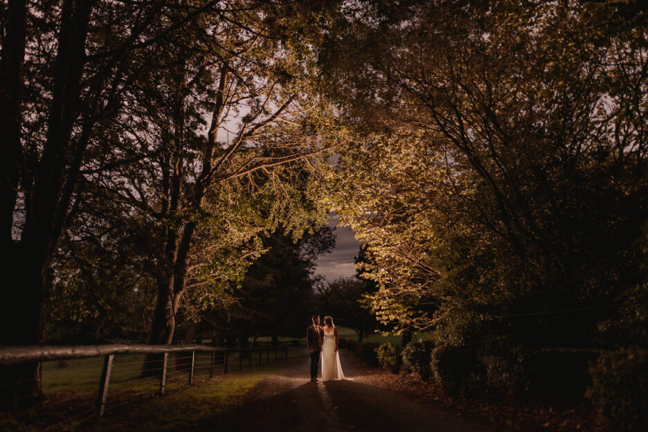 Autumn country back yard wedding bridal couple walking in driveway with trees lit up by flash
