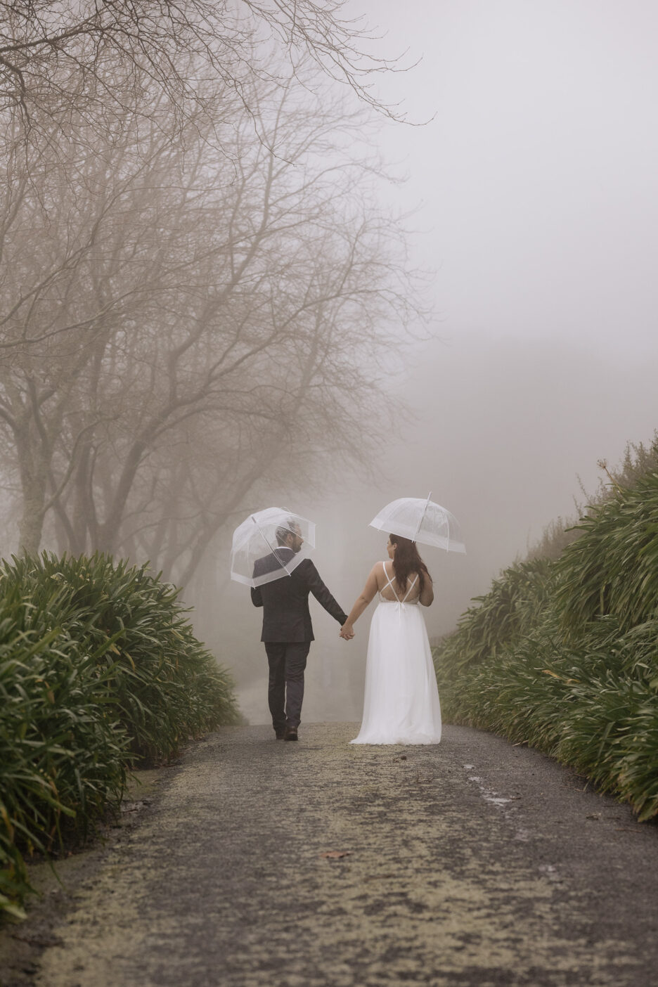 Bride and groom walking away holding hands on driveway in the rain and fog on a winter day