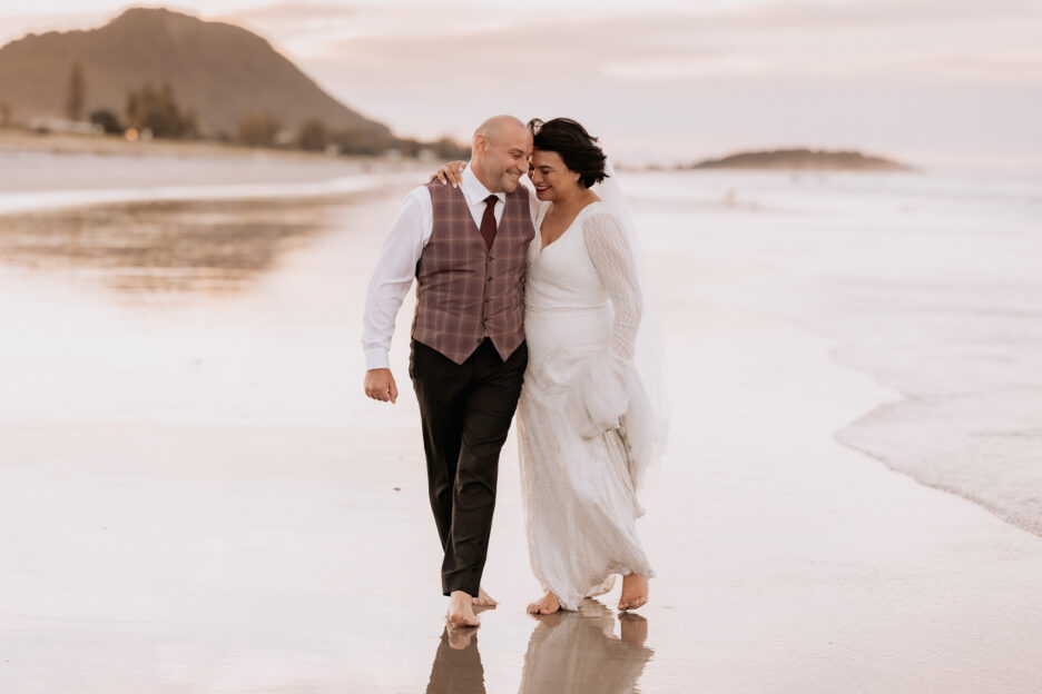 Wedding couple walking on Mount Maunganui beach at dusk, laughing and snuggling together