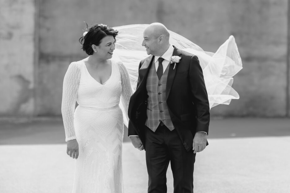 Black and white photo of wedding couple during photoshoot with veil flying behind them, laughing looking at eachother