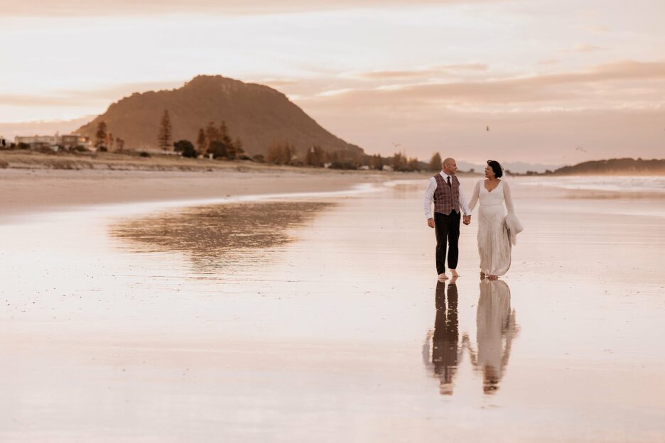 Bride and groom walking at sunset on main mount beach at sunset with mount maunganui behind them
