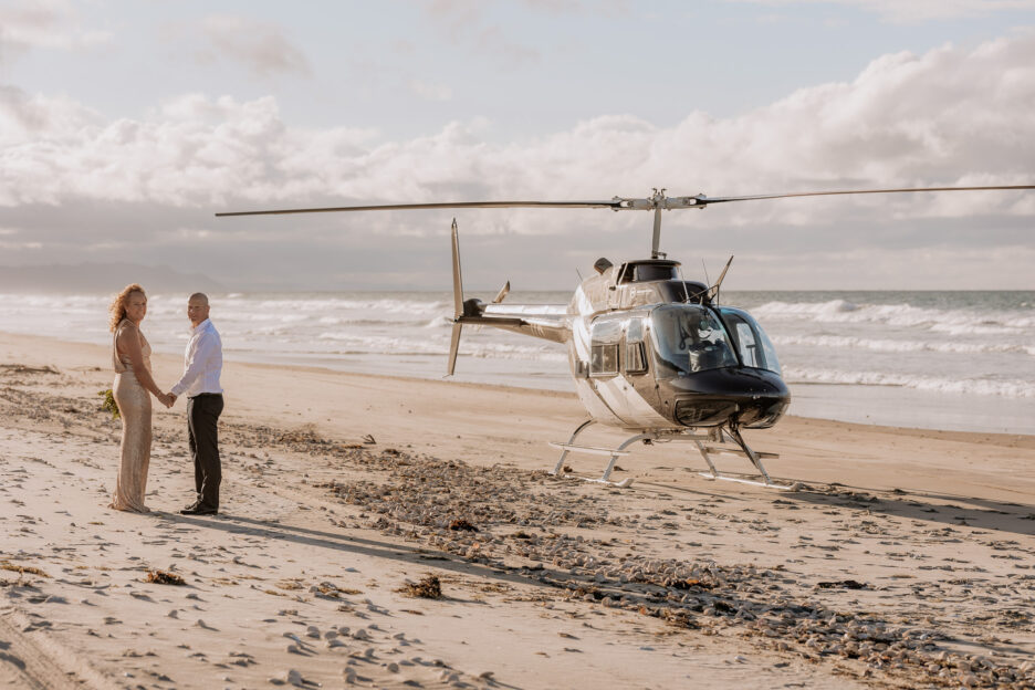 Wedding couple holding hands looking back at photographer beside helicopter on the beach Matakana