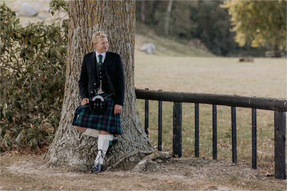 Groom rests against tree, in Scottish kilt