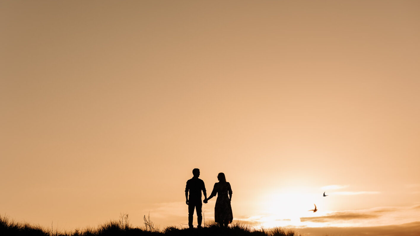 engaged couple standing at sunset Papamoa sand dunes with birds flying