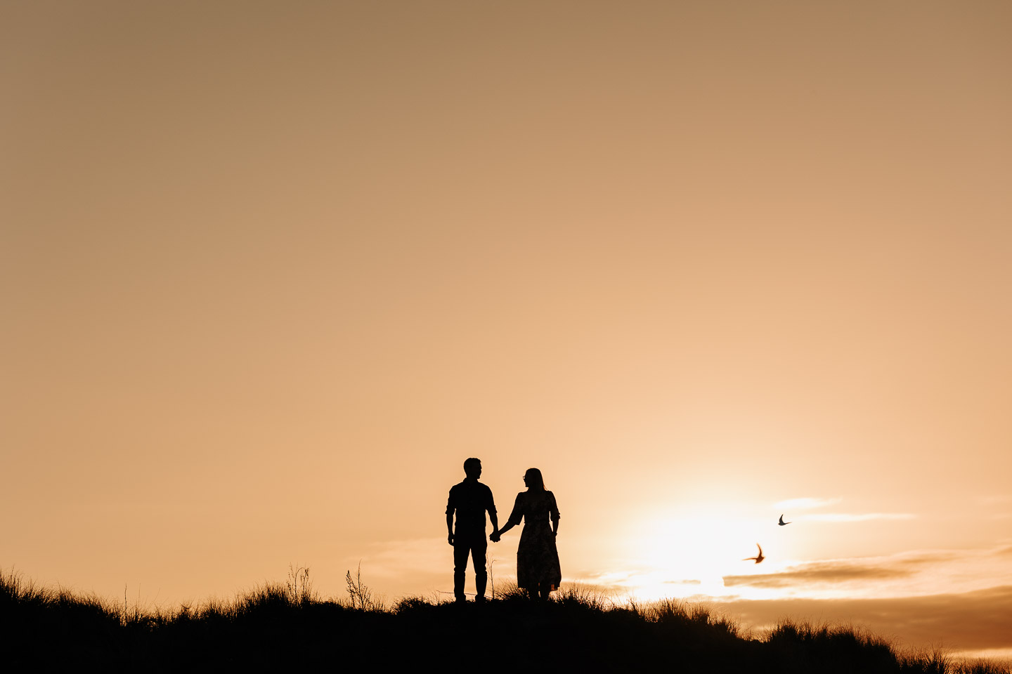 engaged couple standing at sunset Papamoa sand dunes with birds flying