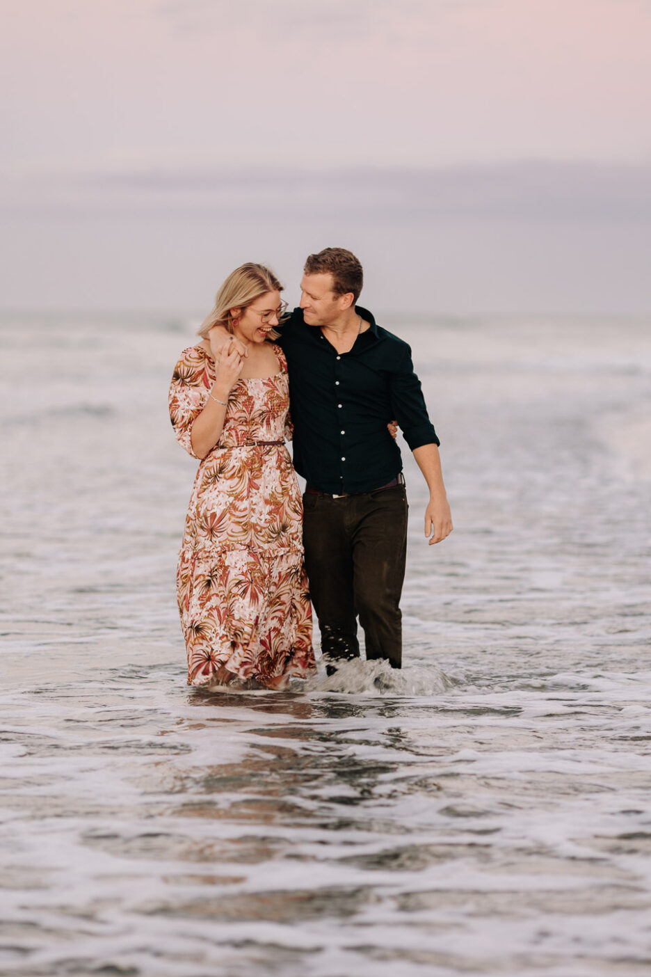 couple get wet walking in the sea on papamoa beach during engagement session