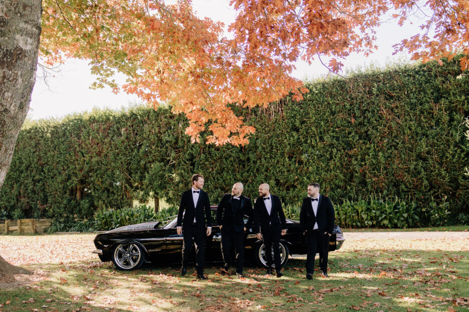 groom with groomsmen in black classic suits standing in front of wedding car under autumn tree