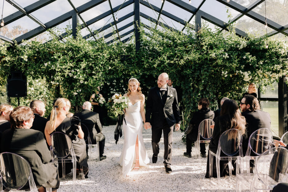 bride and groom walk down aisle under glass house and greenery