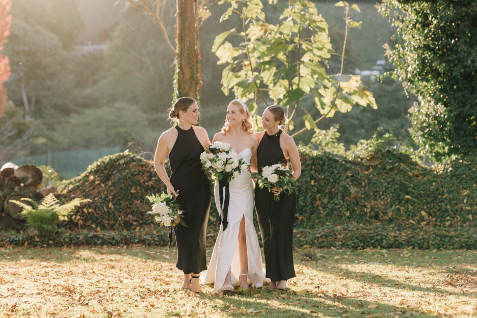 bridesmaids in classic black dresses walk with bride in gardens over looking orchards in Tauranga