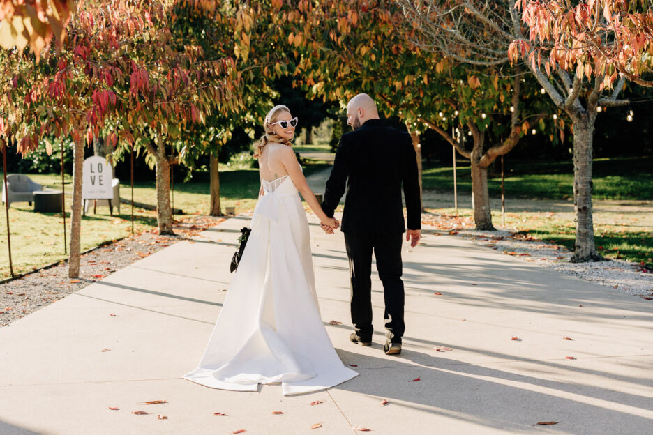 bride with bow on back of dress looks over shoulder as walks away with groom in black suit