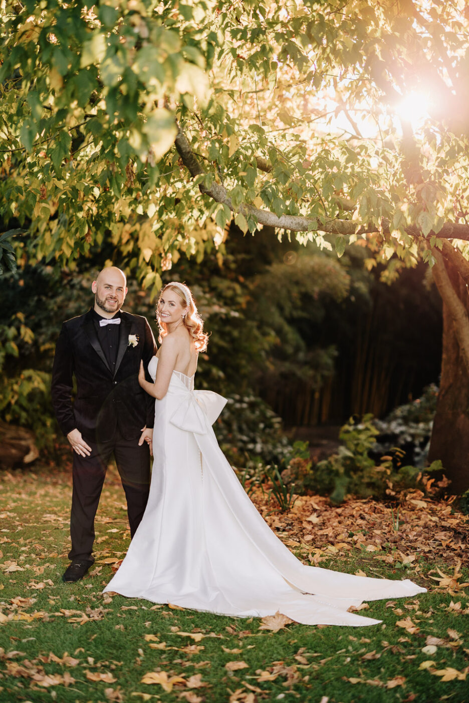 classic photo of bride and groom, bow back of dress, sun comes through tree's behind