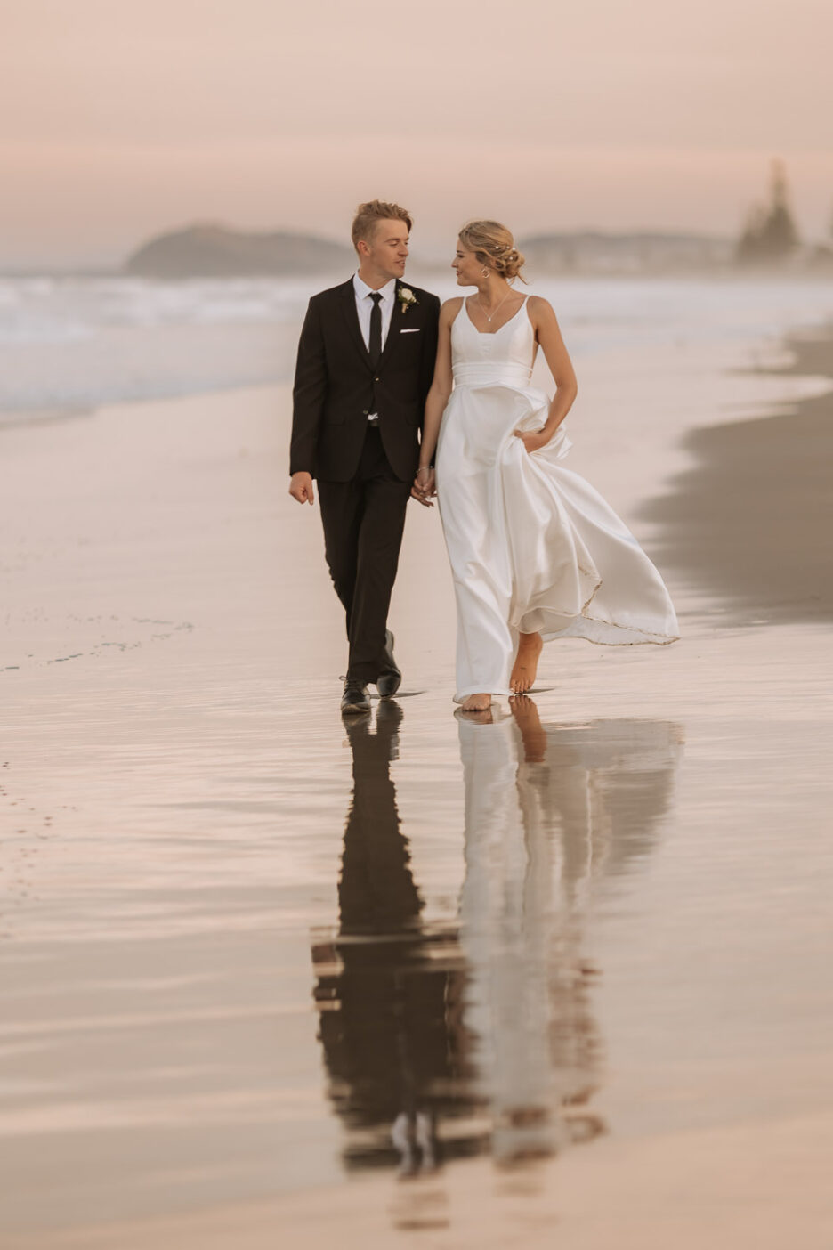 bride and groom walking at golden hour on wet sand at waihi beach