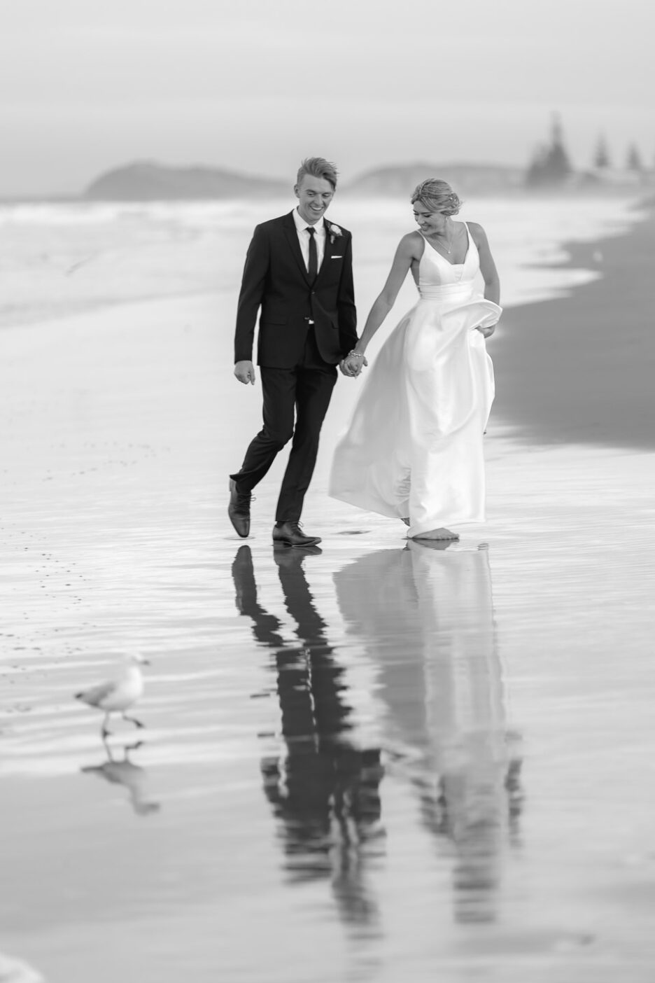 bride and groom walking on wet sand waihi beach wedding photos avoiding waves with seagull