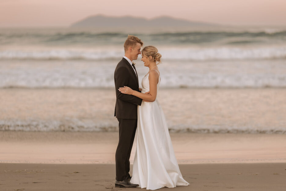 wedding photos in front of waihi beach during golden hour bride and groom standing in front of waves