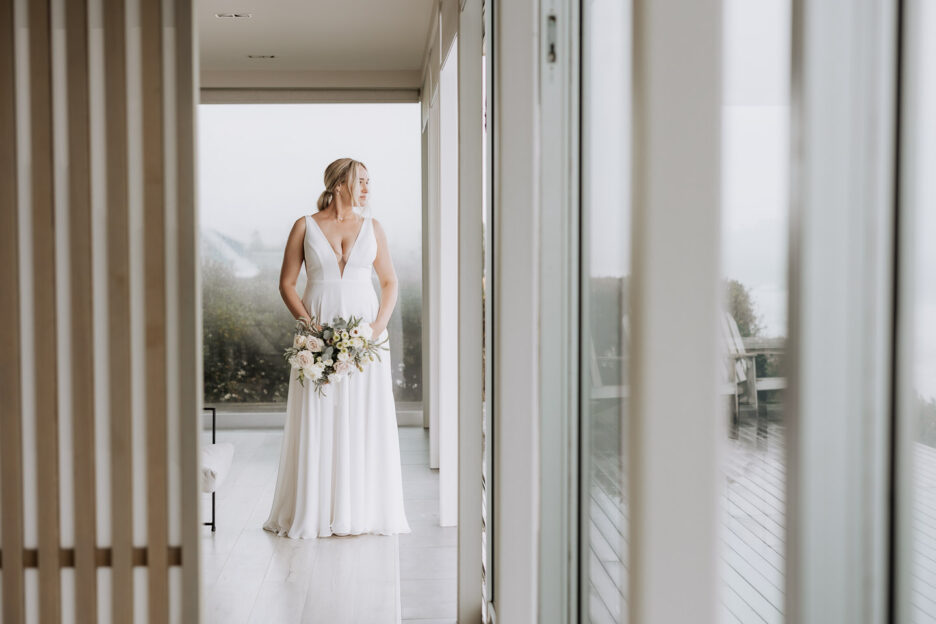 bride standing pukehina beach house holding flowers looking out to sea