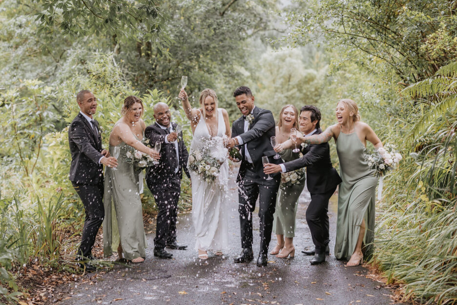 wedding party in green dresses in green driveway of Old forest school opening champagne spray