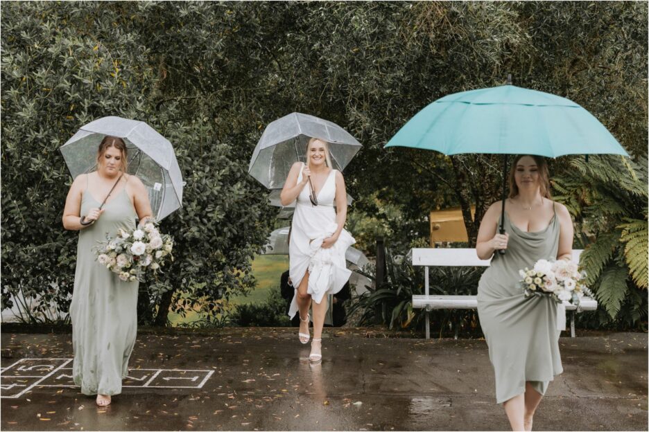 bride walking under umbrellas in the rain on tennis court at old forest school