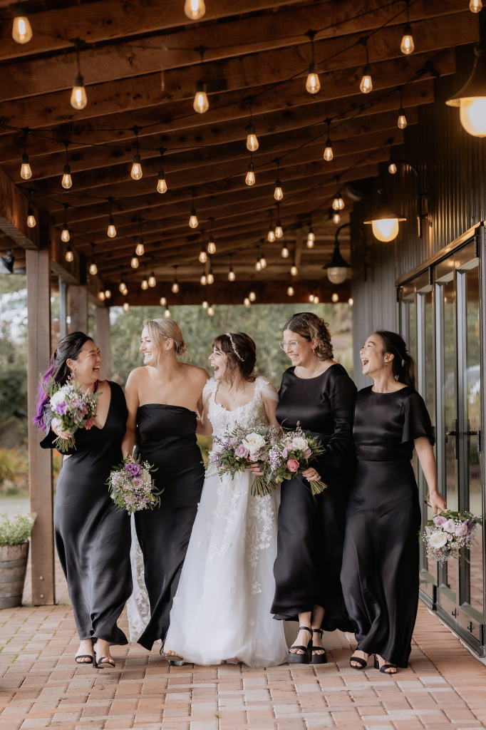 bridesmaids in black dresses laughing and walking under fairy lights